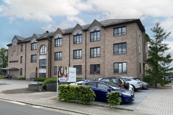 Het gebouw van woonzorgcentrum Berckenbosch, gezien vanaf de straat. Aan de rechterkant van het gebouw bevindt zich een parkeerplaats, terwijl aan de linkerkant een fietsenstalling is. Boven de fietsenstalling is de naam van het woonzorgcentrum zichtbaar op de gevel. Ook is vanuit de straat een bord te zien met de naam en het logo van het centrum, evenals een foto waarop het woord 'welkom' staat.