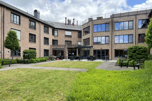 Buitenzicht op het gebouw van woonzorgcentrum Berckenbosch in Heusden. Het pand heeft gevels van lichtrode bakstenen. Er is een terras waar bewoners kunnen zitten op banken of aan tafels. Via verschillende paden is ook de tuin bereikbaar, waar eveneens banken staan om van de omgeving te genieten.