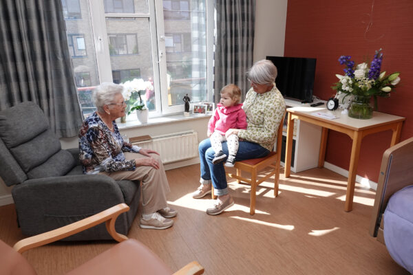 Een eenpersoonskamer in woonzorgcentrum Berckenbosch. Een grootmoeder brengt haar kleindochter, die nog een peuter is, op bezoek bij de overgrootmoeder. De bewoner zit op een grijze zetel, terwijl de bezoekende grootmoeder op een stoel zit en de peuter op haar schoot heeft. De kamer is voorzien van een rode accentmuur en een groot raam, bekleed met grijze gordijnen.