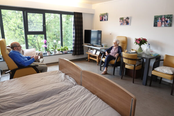 Een dubbele kamer in woonzorgcentrum Berckenbosch in Heusden. De kamer is ingericht met twee naast elkaar geplaatste bedden en bevat vier stoelen, een zwarte tafel, een houten bijzettafel en een tv. Een groot raam zorgt voor veel natuurlijk licht. De twee bewoners zitten tegenover elkaar: de man leest de krant, terwijl de vrouw naar hem kijkt. Aan de muur hangen foto's van hun familie.