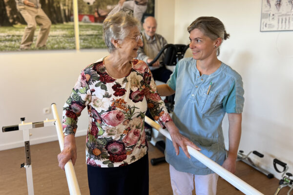 De kinesitherapieruimte van woonzorgcentrum Berckenbosch in Heusden. Een vrouwelijke bewoner, gekleed in een blouse met bloemenmotief, doet wandeloefeningen met behulp van twee leuningen. Een medewerkster ondersteunt haar tijdens de oefeningen, en de bewoner toont blijdschap om de hulp die ze ontvangt.