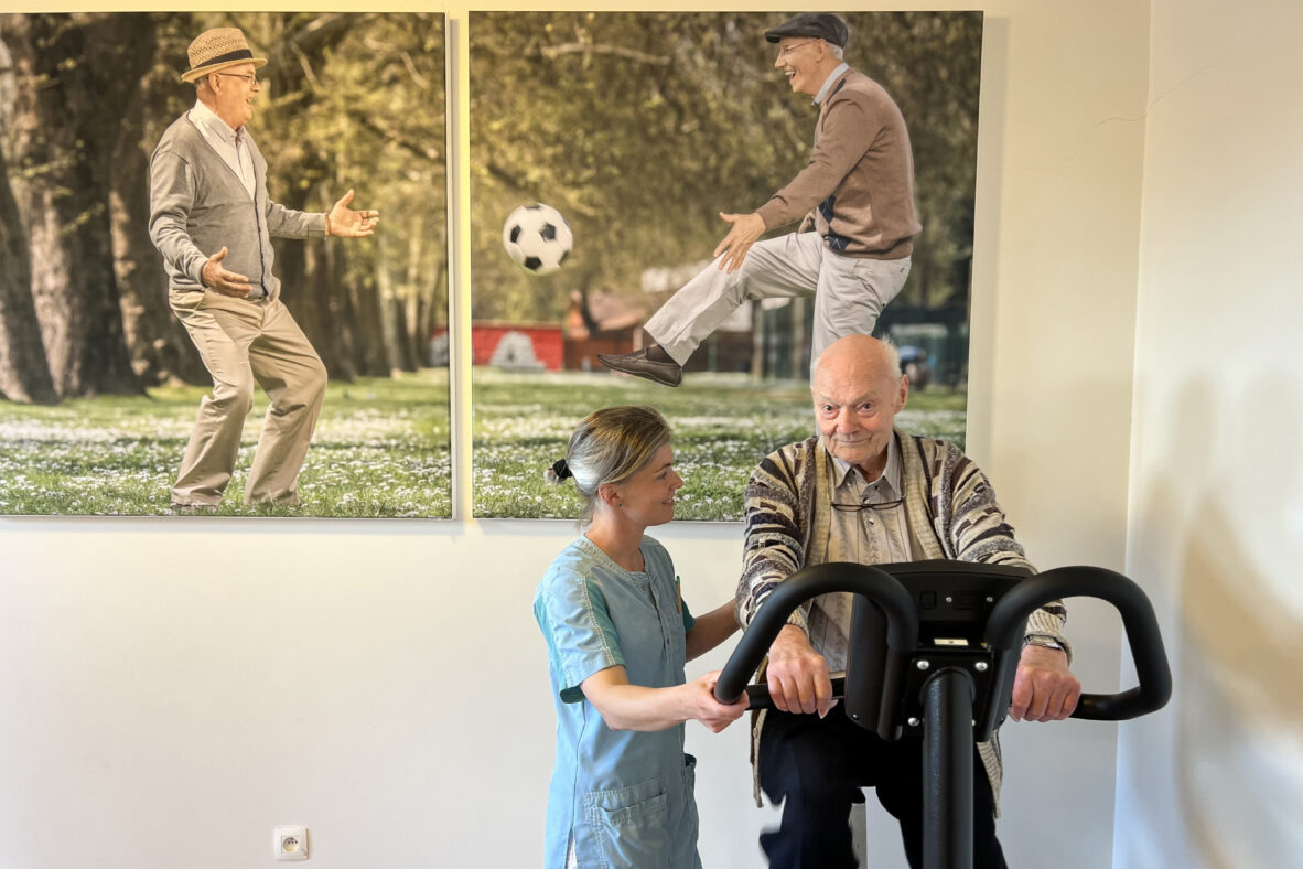 De kinesitherapieruimte in woonzorgcentrum Berckenbosch in Heusden. Een bewoner voert fietsoefeningen uit op een hometrainer, terwijl een medewerkster hem ondersteunt en aanmoedigt. Op de achterwand hangen twee grote foto's die samen een geheel vormen, waarop twee oudere mannen te zien zijn. Eén man schopt een voetbal naar de andere.