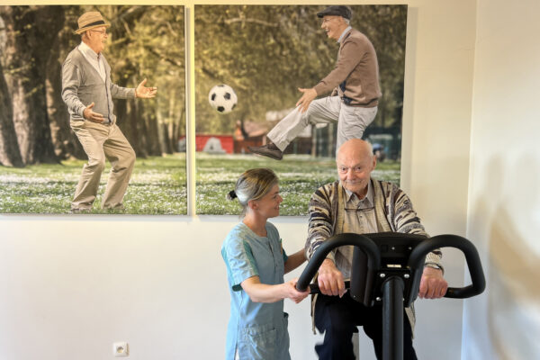 De kinesitherapieruimte in woonzorgcentrum Berckenbosch in Heusden. Een bewoner voert fietsoefeningen uit op een hometrainer, terwijl een medewerkster hem ondersteunt en aanmoedigt. Op de achterwand hangen twee grote foto's die samen een geheel vormen, waarop twee oudere mannen te zien zijn. Eén man schopt een voetbal naar de andere.