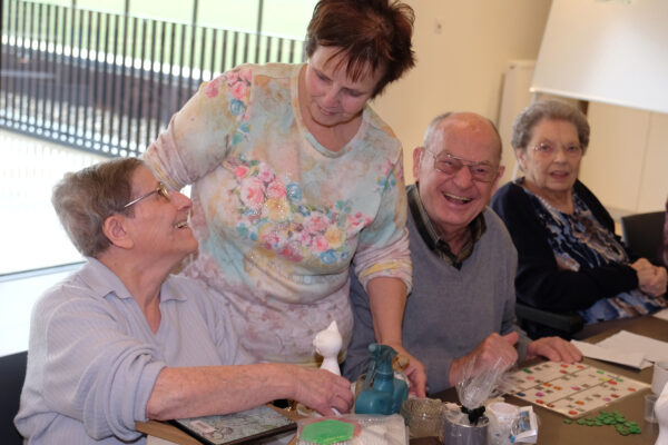 Drie bewoners van woonzorgcentrum Berkenhof in Heers spelen samen spelletjes en lachen. Achter hen staat een vrijwilliger die met een van de bewoners in gesprek is over een klein porseleinen standbeeld van een kat.