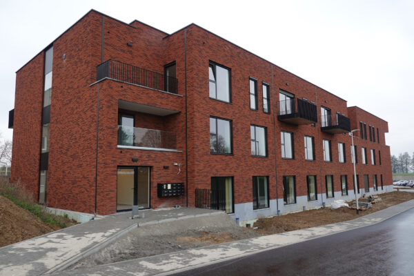 Het uitzicht vanuit de straat op het gebouw van de assistentiewoningen Berkenhof in Heers toont een gevel van baksteen in verschillende tinten rood. Een pad leidt naar de glazen voordeur met een zwart kozijn. Naast de deur bevinden zich brievenbussen voor elke flat. Op de voorgevel zijn zes balkons zichtbaar: twee uitspringende balkons en vier ingebouwde balkons. Sommige terrassen hebben zwarte metalen balustrades, terwijl andere voorzien zijn van glazen leuningen.