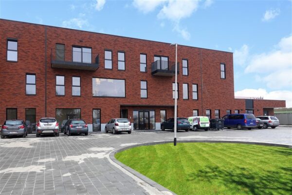 Zicht op de buitengevel en de parking van woonzorgcampus Berkenhof. Negen auto's staan geparkeerd op de parkeerplaats. De gevel is opgebouwd uit baksteen in verschillende rode tinten. Op de tweede verdieping bevinden zich twee terrassen die verschillen van grootte.