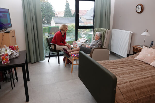 Een bewoner van woonzorgcentrum Berkenhof ontvangt bezoek in haar privatieve kamer. Het bed is netjes opgemaakt, met twee sierlijke kussens. De bewoner zit ontspannen in haar lichtbruine zetel, haar benen rustend op een voetstoel. De bezoeker zit naast haar op een zwarte stoel en voert een gesprek met haar. Op de bijzettafel naast hen liggen verschillende wenskaarten en een cadeaumandje. De tv, die op een kast staat, is aan. Een groot raam biedt uitzicht op de tuin en kan worden bedekt met groene gordijnen.