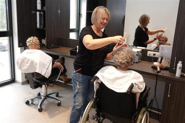 De kapster van woonzorgcampus Berkenhof heeft twee bewoners in haar kapsalon. Ze kamt het haar van een bewoner, terwijl de andere voor de spiegel zit met haarrollers in haar haar.