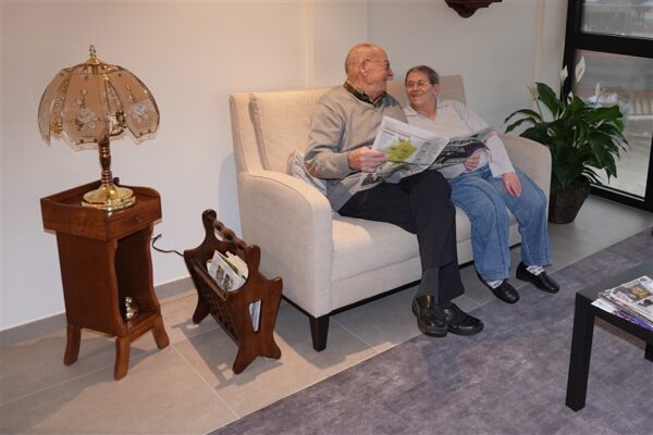 Een mannelijke en vrouwelijke bewoner van de assistentiewoningen Berkenhof zitten op een beige sofa in het ontspanningssalon. Ze kijken naar elkaar en glimlachen. Naast hen staat een oud houten bijzettafeltje met een decoratieve roze lamp erop.