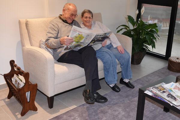 Twee bewoners van woonzorgcentrum Berkenhof zitten samen op een beige sofa in de ontspanningsruimte. Ze lezen gezamenlijk een Limburgse krant. Op de betegelde vloer ligt een grijs tapijt.
