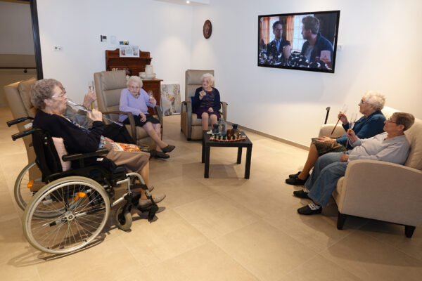 Zes nieuwe bewoners van het woonzorgcentrum Berkenhof in Heers worden hartelijk verwelkomd en zitten samen in het ontspanningssalon. Drie van hen nemen plaats in eenpersoonszetels, terwijl twee anderen gezellig samen op de sofa zitten. De laatste bewoner zit comfortabel in haar rolstoel. Gezamenlijk genieten ze van een glas champagne en voeren een ontspannen gesprek. Aan de muur hangt een televisie die op dat moment aanstaat.