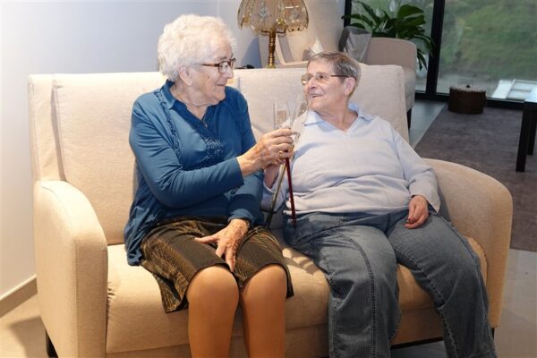 Twee bewoners van de assistentiewoningen Berkenhof zitten in het gemeenschappelijke ontspanningssalon en drinken iets. Ze zitten op een beige sofa en kijken naar elkaar.