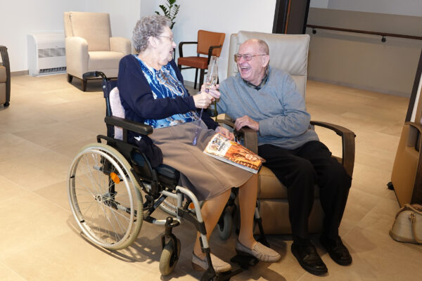 Twee bewoners van woonzorgcentrum Berkenhof zitten in de gemeenschappelijke ontspanningsruimte. De mannelijke bewoner zit op een stoel, terwijl de vrouwelijke bewoner in haar rolstoel zit. Ze lachen samen en drinken een drankje terwijl ze proosten.