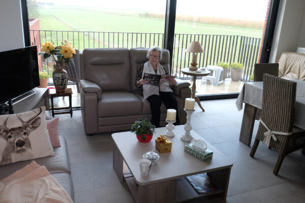 Een bewoner van de assistentiewoningen in Berkenhof zit op een grijze sofa in haar woonkamer, verdiept in een magazine. De woonkamer biedt toegang tot een groot terras via een schuifdeur. Daarnaast staat er een tweede sofa en een salontafel, waarop twee kaarsen en een bloemenpot ter decoratie staan.