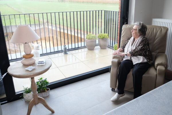 Een bewoner van een serviceflat in woonzorgcampus Berkenhof zit in haar beige fauteuil en kijkt door het raam naar buiten. De grote schuifdeur biedt toegang tot een terras met beige tegels en een zwarte metalen leuning. Op het terras staan standbeelden van een hond en een kat.