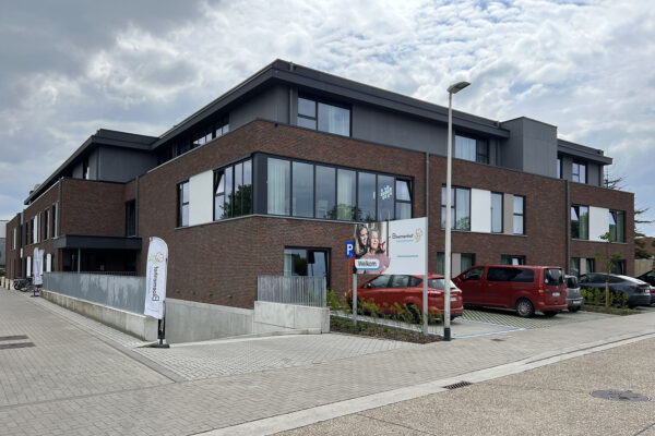 Uitzicht vanaf de straat op het gebouw van woonzorgcentrum Bloemenhof. Langs de straat is parkeerruimte beschikbaar, en er is tevens toegang tot een ondergrondse parking. Een bord met een foto van een oudere vrouw en haar familielid, samen met de naam en het logo van het woonzorgcentrum, verwelkomt de bezoekers.