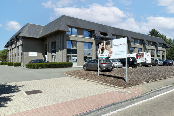 Straatzicht op het gebouw van de assistentiewoningen Bruggenpark. Voordat men de parking betreedt, komt men langs een bord met een foto, de naam en het logo van het woonzorgcentrum. Het bord vermeldt ook dat de woonzorgcampus zowel assistentiewoningen, een woonzorgcentrum als een hersteverblijf biedt.