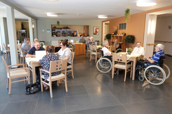 De brasserie van woonzorgcentrum Bruggenpark. Achteraan staan twee vrijwilligers achter de bar en serveren de bestellingen. Meerdere bewoners zitten samen aan tafels en voeren gesprekken met elkaar. Andere bewoners zijn in gezelschap van een familielid dat op bezoek is. De vloer is betegeld met grijze tegels, en de muren zijn versierd met behang in verschillende kleuren, waaronder oranje en lichtgroen.