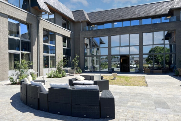 Zicht op de achterkant van het gebouw van woonzorgcentrum Bruggenpark. Het gebouw heeft donkergrijze bakstenen gevels en een dak bedekt met donkergrijze shingles. De bovenste verdieping beschikt over een groot terras met glazen leuningen, dat uitzicht biedt op de tuin.