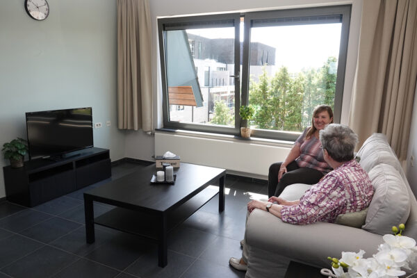 Een bewoner van de assistentiewoningen Bruggenpark zit samen met haar dochter op een witte sofa in de woonkamer van haar flat. De ruimte beschikt over twee grote ramen, waarvan één in een knikvorm openstaat, waardoor veel natuurlijk licht naar binnen komt. De ramen kunnen worden afgedekt met beige gordijnen.