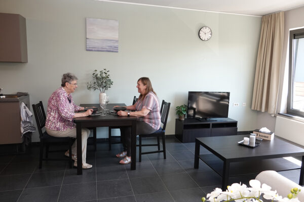 Een betegelde flat in de assistentiewoningen van Bruggenpark. In de zitruimte staat een witte sofa, een zwarte salontafel en een tv. De bewoner zit samen met haar dochter, die op bezoek is, in de eetkamer. Deze ruimte is ingericht met een zwarte rechthoekige tafel en vier bijpassende stoelen, en aan de muur hangt een schilderij van de zee.