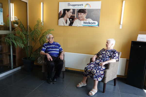De inkomhal van woonzorgcentrum Bruggenpark, met geel geschilderde muren waarop een plakkaat hangt dat bezoekers verwelkomt. In de hal staan twee stoelen, waarop twee bewoners zitten.