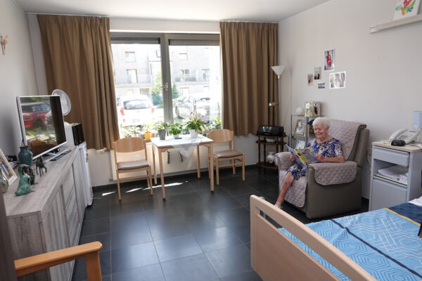 Een privatieve kamer in woonzorgcentrum Bruggenpark, ingericht met een bed, een zetel, een tafel met twee stoelen, een kleine koelkast, een magnetron, een bijzettafel met een radio, een nachtkastje met een telefoon en een commode met een televisie erop. De bewoner zit op de zetel en leest rustig een tijdschrift.