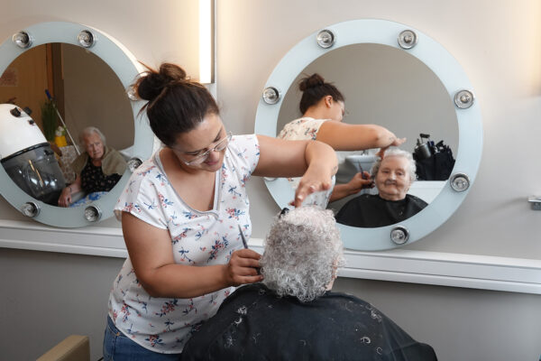 Detailfoto van het kapsalon in woonzorgcentrum Bruggenpark. De kapper knipt het gekrulde haar van een bewoner, die zichzelf in de spiegel kan zien tijdens haar knipbeurt.