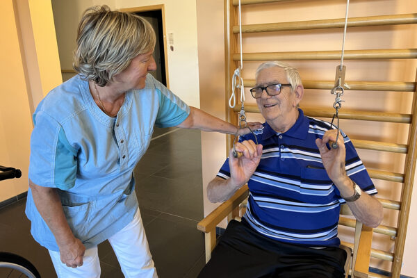 Binnen in de kinesitherapieruimte van woonzorgcentrum Bruggenpark. Een mannelijke bewoner zit op een stoel met zijn rug tegen een klimtoestel waaraan een fitnesselastiek is bevestigd. Hij gebruikt het elastiek voor armoefeningen, terwijl een medewerkster naast hem staat en hem ondersteunt.