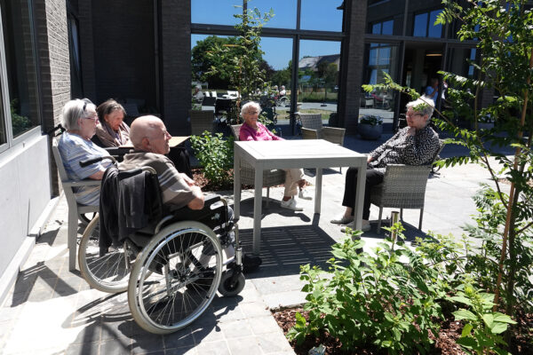 Vijf bewoners zitten rondom een houten tafel op een zonnige dag op het terras van woonzorgcentrum Bruggenpark. Drie bewoners zitten op rieten stoelen, terwijl de andere twee in hun rolstoelen zitten. Het terras is betegeld met grijze tegels en is voorzien van planten en bomen.