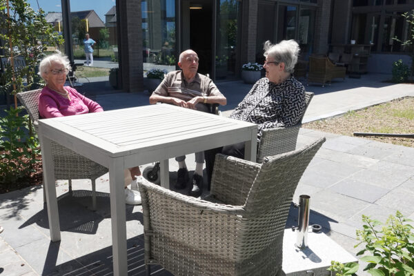 Drie bewoners zitten samen in de zon aan een houten tafel op het terras van woonzorgcentrum Bruggenpark. De tafel heeft nog een vierde stoel. Via het terras is er een deur die toegang biedt tot het gebouw.