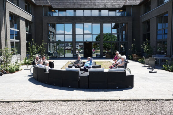 Negen bewoners zitten op het terras van woonzorgcentrum Bruggenpark. Het ruime terras is uitgerust met twee banken: een tweepersoonsbank en een grote, halfcirkelvormige bank die plaats biedt aan zes personen. Op de achtergrond is het gebouw van het woonzorgcentrum zichtbaar, met grote ramen.