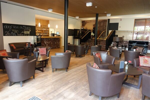 Een overzichtsfoto van de brasserie in woonzorgcampus Clarenhof. Op een krijtbord staat aangegeven dat er op vrijdag 26 mei een barbecue zal plaatsvinden. De brasserie heeft een parketvloer, terwijl de trappen en de bar zijn gemaakt van marmer met goudkleurige accenten.