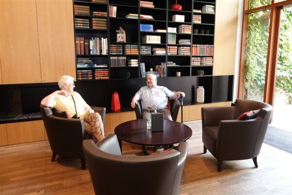 De brasserie van woonzorgcampus Clarenhof. Twee bewoners zitten op bruine lederen fauteuils en voeren een gesprek rond een ronde, bruine bijzettafel. Achter hen staat een grote boekenkast, gevuld met boeken.