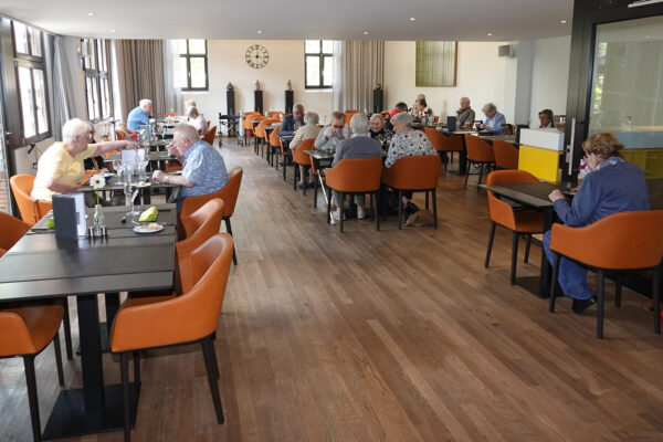 De brasserie van woonzorgcentrum Clarenhof, waar verschillende mensen aan het eten of drinken zijn. De ruimte heeft een parketvloer en de wanden zijn wit geschilderd. De tafels zijn zwart, terwijl de stoelen oranje zijn met zwarte poten. Sommige mensen eten alleen, anderen in groepen.