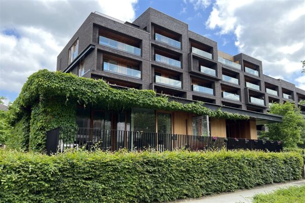 Straatzicht op de assistentiewoningen van Clarenhof. De ingebouwde balkons op de eerste tot en met de vierde verdieping zijn voorzien van glazen leuningen. De terrassen op de begane grond hebben zwarte verticale metalen leuningen. Aan de gevel van het gebouw groeit een klimplant omhoog.