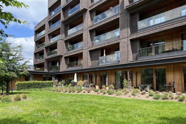 Het gebouw van de assistentiewoningen Clarenhof, gezien vanaf de achterkant. De gevel is voornamelijk opgebouwd uit grijze bakstenen, met hier en daar houten accenten bij de terrassen. Alle terrassen beschikken over grote schuifdeuren, en de terrassen op de hogere verdiepingen hebben glazen leuningen.