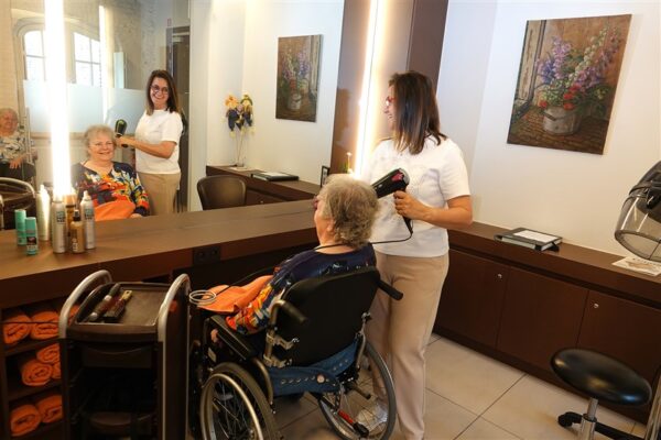 De kapper van woonzorgcampus Clarenhof droogt het haar van een bewoner, die glimlachend naar zichzelf kijkt in de spiegel. De bewoner zit in haar rolstoel. Aan de muur van het kapsalon hangt een schilderij van verschillende bloemen.