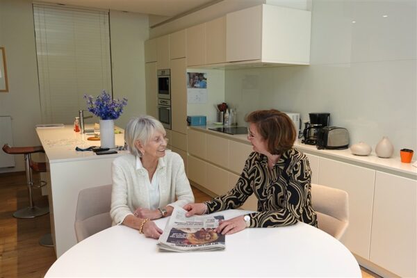 Een bewoner van de assistentiewoningen van Clarenhof ontvangt bezoek in haar serviceflat. Ze zitten samen aan een ronde witte tafel in de eetkamer en bekijken de krant. Achter hen is de keuken zichtbaar, met zijn strakke witte kasten.