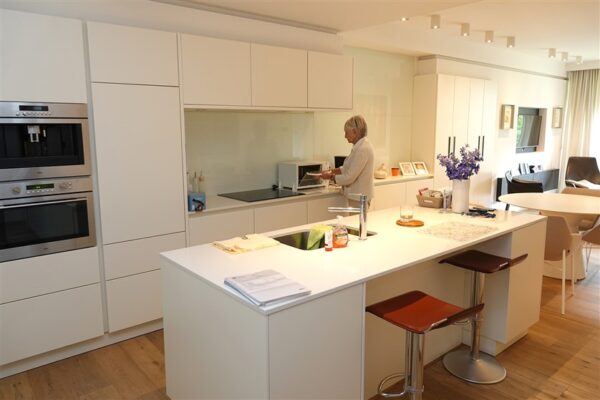 Een moderne, witte keuken in een van de assistentiewoningen van Clarenhof. In het midden staat een groot kookeiland met twee barstoelen. De kraan en de spoelbak bevinden zich op het eiland. De keuken is voorzien van een ingebouwde magnetron en oven, die boven elkaar zijn geplaatst. Vanuit de keuken zijn de eet- en zitkamer zichtbaar.