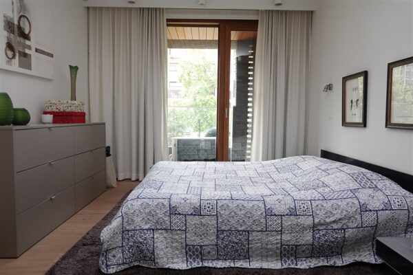 De slaapkamer van een serviceflat in Clarenhof is stijlvol ingericht. In het midden van de kamer staat een opgemaakt bed, tegenover een grijze ladekast. De muren zijn versierd met diverse schilderijen en vanuit de slaapkamer is er toegang tot een terras.