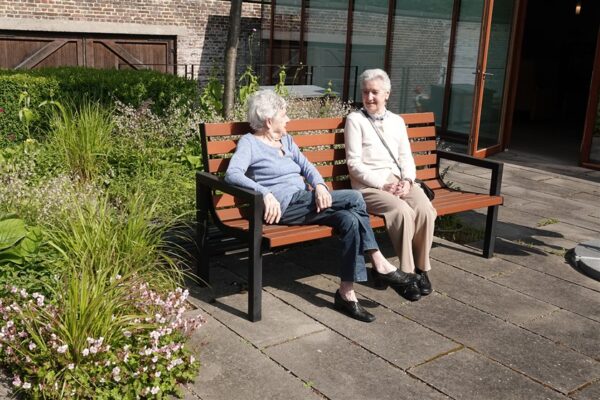 Twee bewoners van woonzorgcentrum Clarenhof genieten samen van de zon op een bruine bank in de tuin. Ze worden omringd door weelderige planten, terwijl achter hen een deur zichtbaar is die toegang biedt tot het gebouw van het woonzorgcentrum.