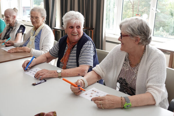 Meerdere bewoners van woonzorgcentrum Coham nemen deel aan een potje bingo. Ze zitten naast elkaar aan een tafel, elk met een stift in een andere kleur.