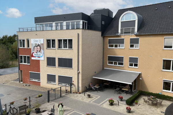 Bovenaanzicht op het terras van woonzorgcentrum Coham. Op het terras, die zowel langs het gebouw als de straat betreedbaar is, zitten twee bewoners rustig iets te drinken. Op de gevel van het gebouw hangt een bord met een foto van een oudere vrouw en een dierbare van haar die haar omhelsd.