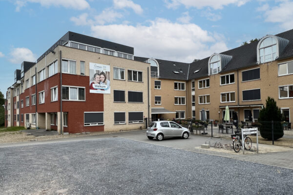 Uitzicht op het gebouw van woonzorgcentrum Coham. De gevels zijn opgebouwd uit bakstenen in drie verschillende kleuren: rood, lichtbeige en lichtoranje. Aan de gevel hangt een groot bord met de naam en het logo van het woonzorgcentrum, waarop in grote letters het woord "Welkom" is geschreven.