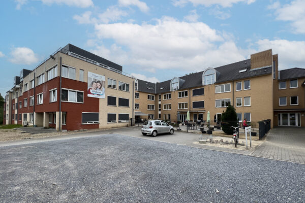 Uitzicht op het gebouw, het buitenterras en de parkeerplaatsen van woonzorgcentrum Coham. De ingang van het gebouw bevindt zich aan de rechterkant, naast een fietsenstalling. Op de parkeerplaats staat een grijze auto geparkeerd, een fiets is in de fietsenstalling geplaatst, en een persoon wandelt op het terras.