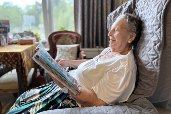 Een bewoonster van woonzorgcentrum Coham zit op een fauteuil in haar kamer en leest een stripverhaal. Op de achtergrond is een tafel en een antieke stoel zichtbaar met daarop een kussen die een bloemenmotief heeft.