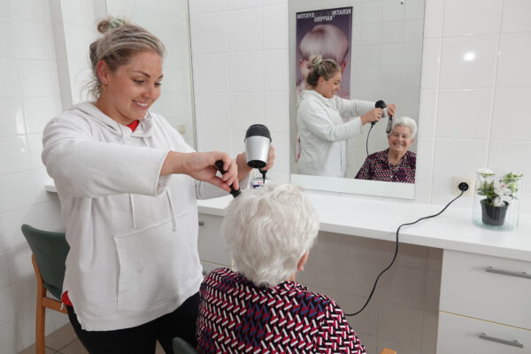 De kapster van woonzorgcentrum Coham kamt en droogt het haar van een bewoonster. Op de muur van de kapsalon, die betegeld is met witte tegels, hangt een spiegel waarin de bewoonster zichzelf kan zien.