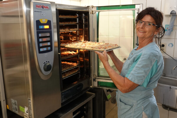Een medewerkster van woonzorgcentrum Coham, die een bril draagt, zit in de keuken en plaatst meerdere pizza's in een grote oven die op 180 graden is voorverwarmd.