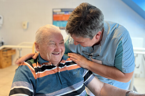 Een bewoner van woonzorgcentrum Coham zit in de kinéruimte. Een medewerkster houdt hem vast en zegt iets wat hem doet glimlachen.